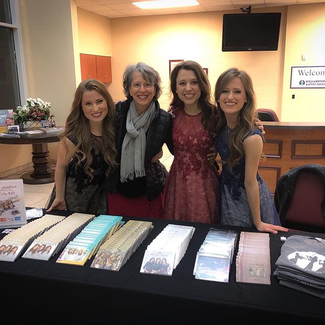 Three daughters stand with their mother behind a merchandise table with CDs and custom t-shirts