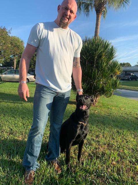 A man stands in a yard smiling with his hand on the head of his emotional support dog.