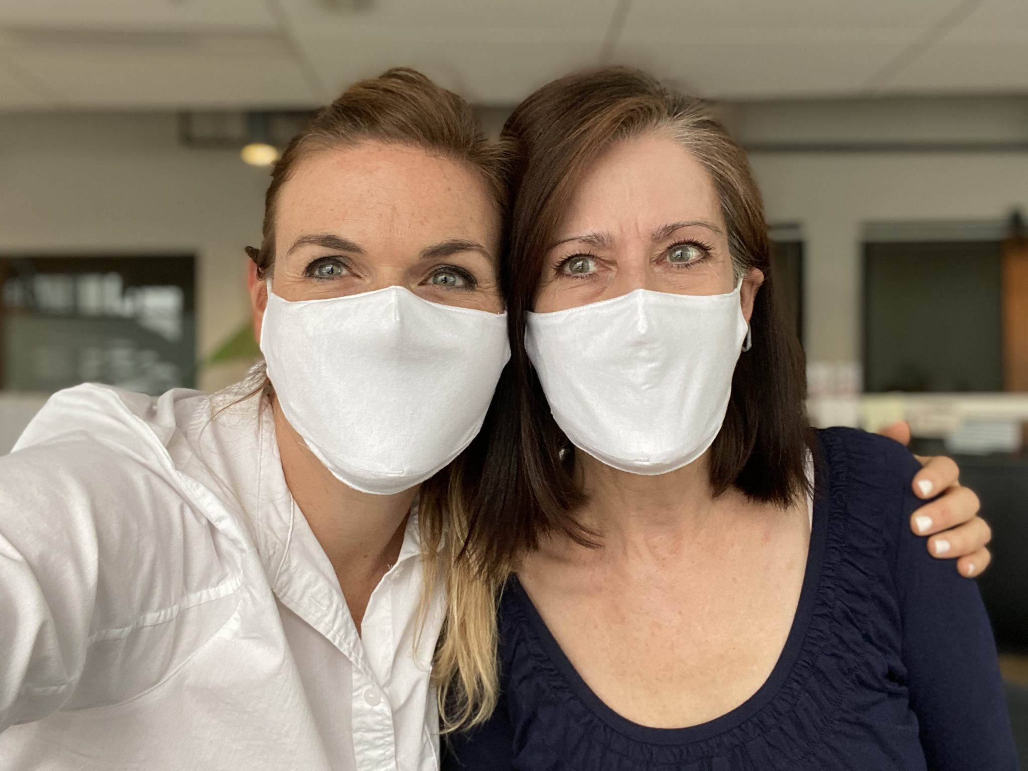 Two women smiling wearing white masks. One has her arm around the other one.