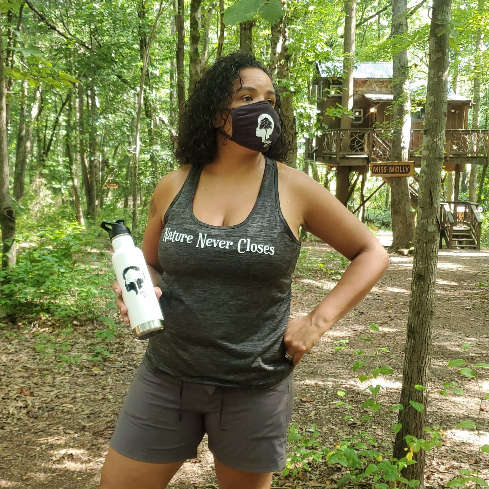 A woman stands in the woods wearing a custom face mask and custom water bottle both with the Outdoor Afro logo (a person's silhouetted head with a tree growing inside it) and a custom tank top that says "Nature Never Closes."
