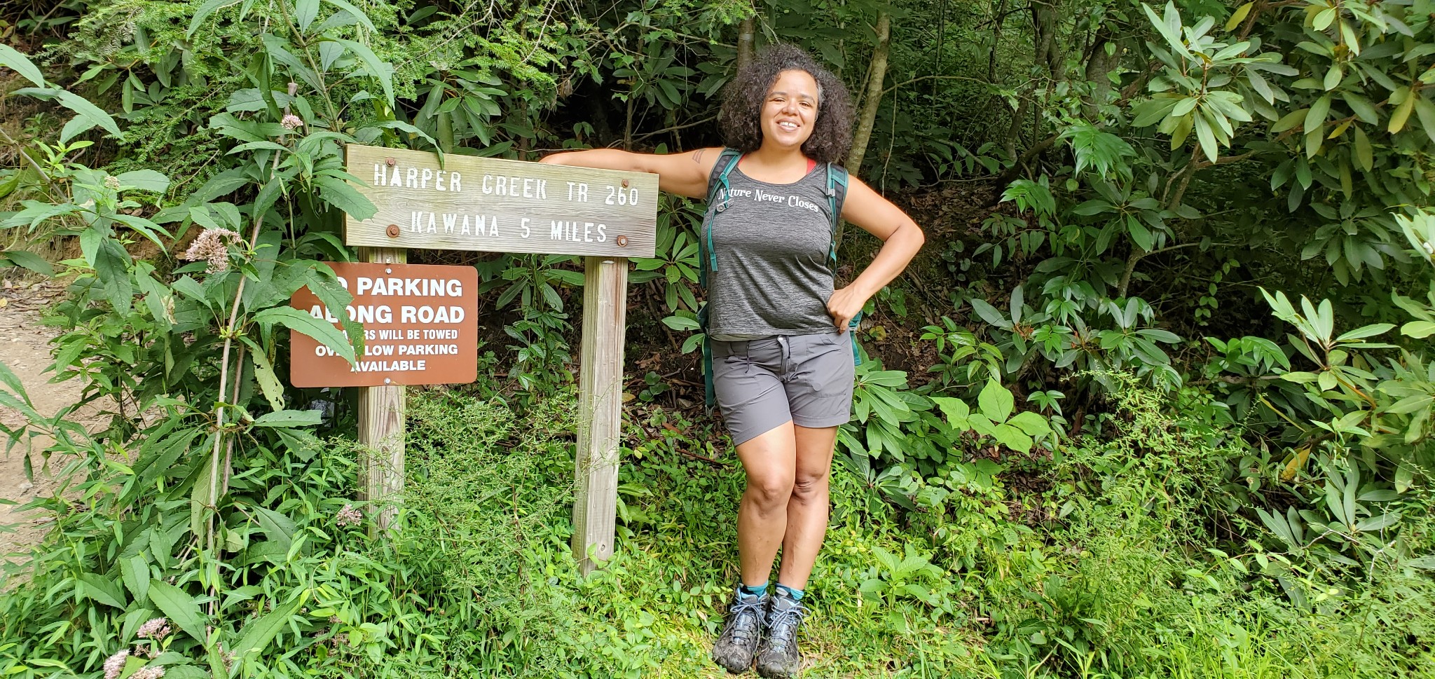 A woman stands in the woods surrounded by trees with her arm around a hiking trail sign. She's smiling and wearing a custom tank top that says "Nature Never Closes."