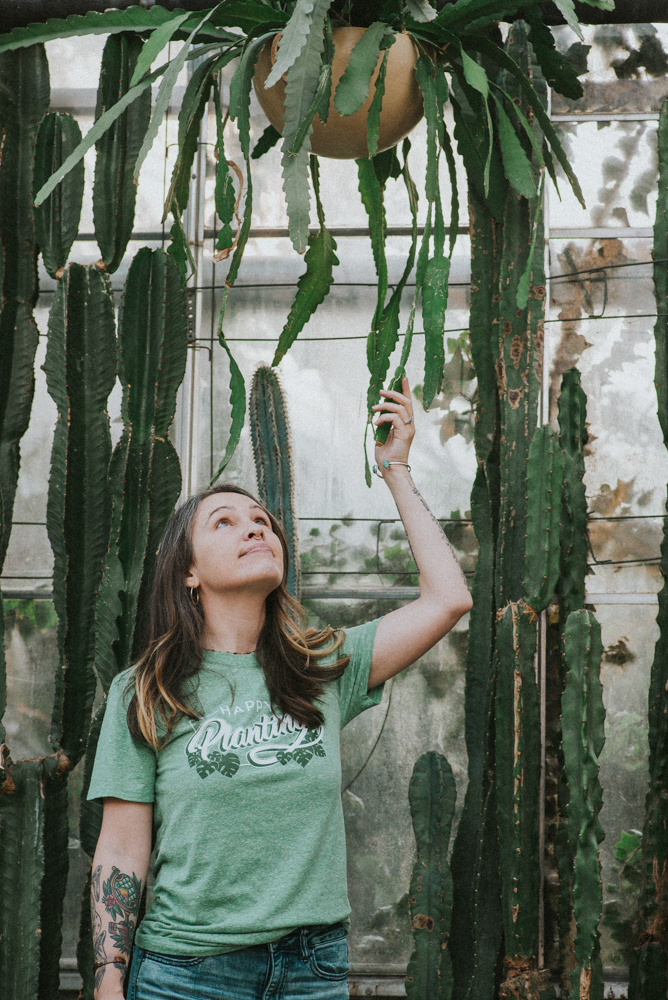 A woman with long brown hair wearing a green custom t-shirt that says "Happy Planting" looks up at and touches a hanging plant.