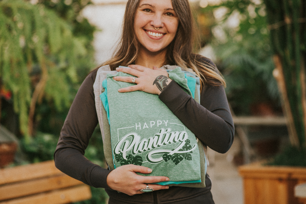 A smiling woman holds folded green custom shirts with the words "Happy Planting" on them to her chest.