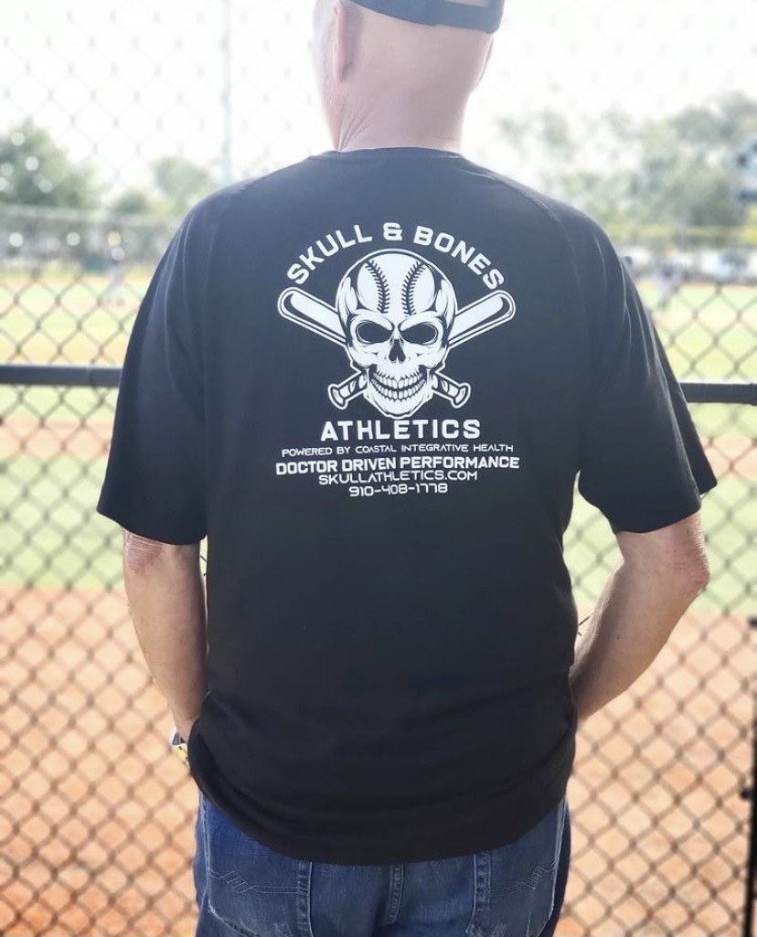 Back of man at a baseball field wearing a shirt with the text Skull & Bone Athletics printed on it