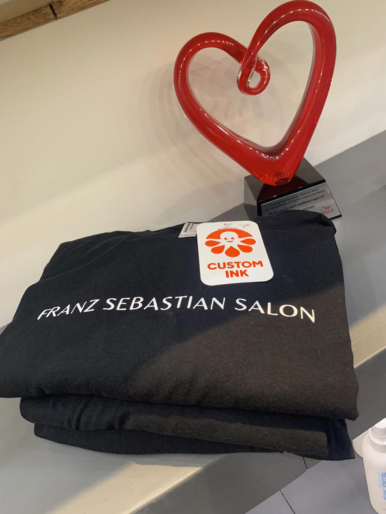 A stack of custom black t-shirts with the text Franz Sebastian Salon in white printed on them.
