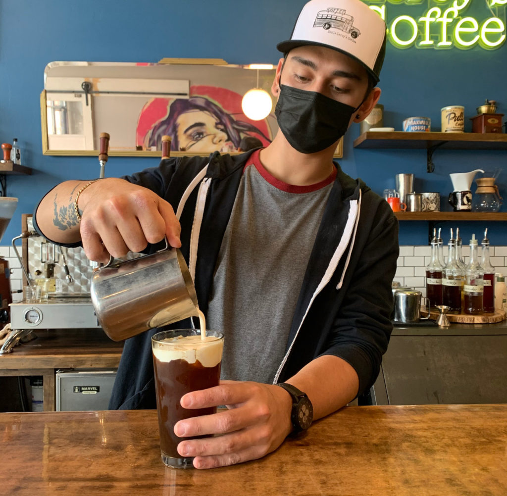 Man wearing Uncle Leroy's Coffee hat makes a coffee drink in a cafe