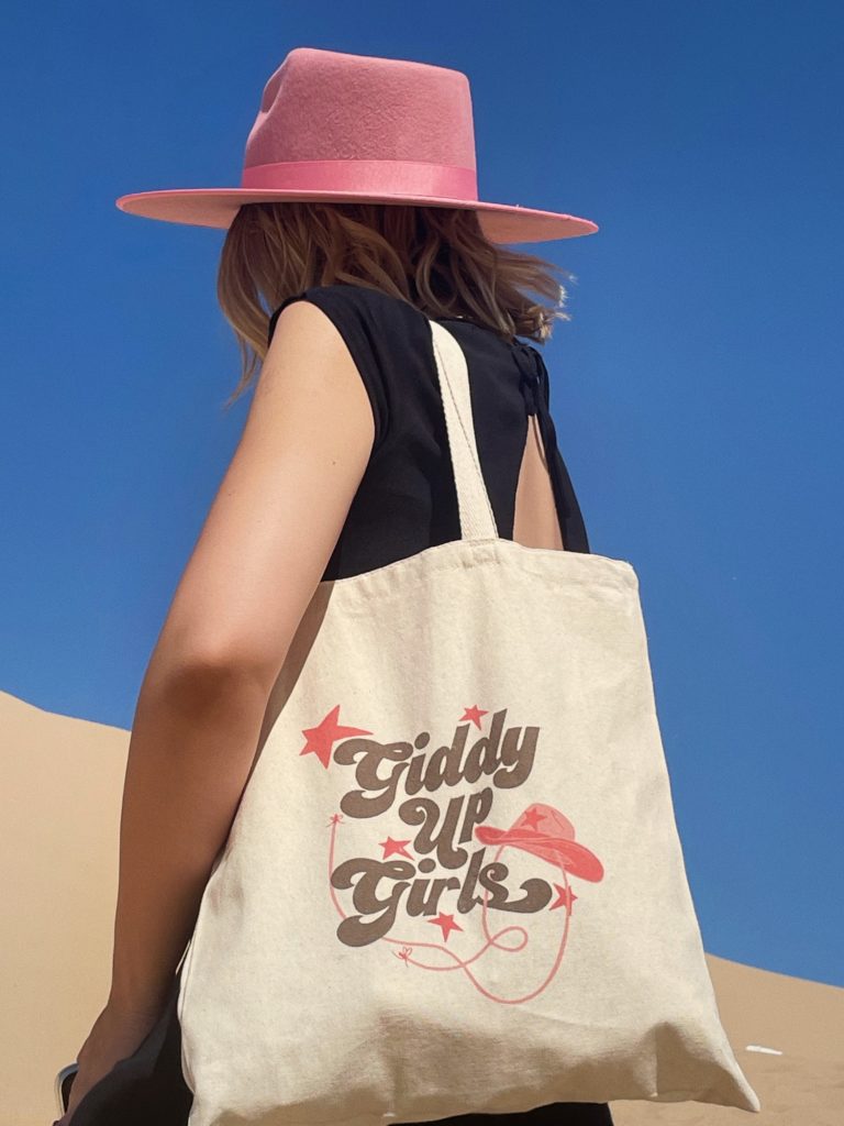 Woman wearing pink cowboy hat shows off tote bag with Giddy Up Girls printed on the side