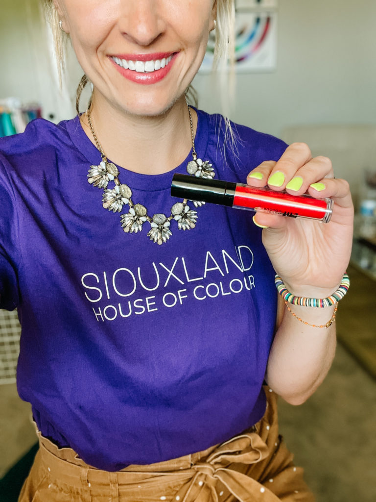 Close-up of woman holding lip gloss and wearing a purple t-shirt with Siouxland House of Color printed on it.