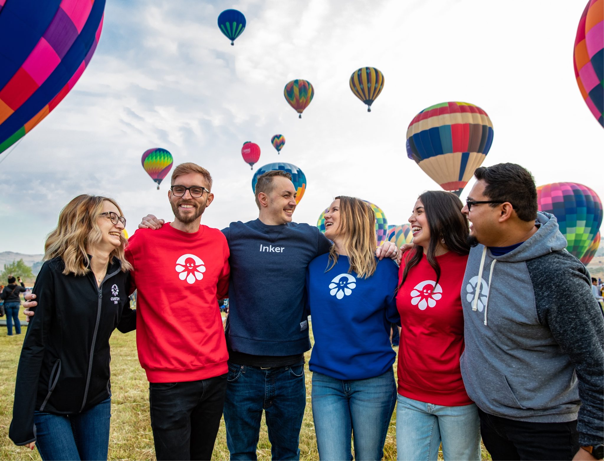 Six people stand in front of hot air balloons. They're wearing Custom Ink sweatshirts and t-shirts featuring the CI mascot, Inky