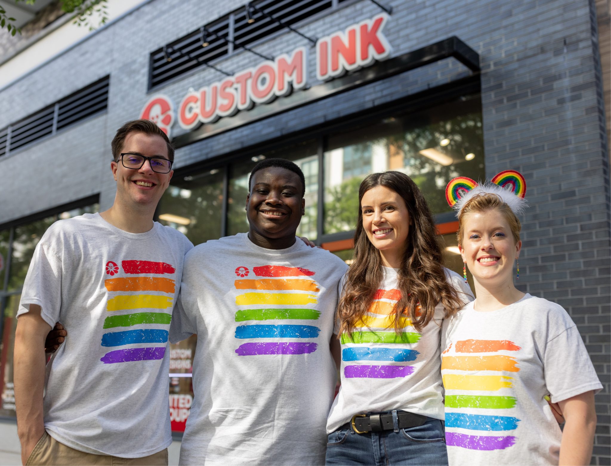 Four people stand in front of a Custom Ink store. They are wearing t-shirts with rainbow designs on the front.