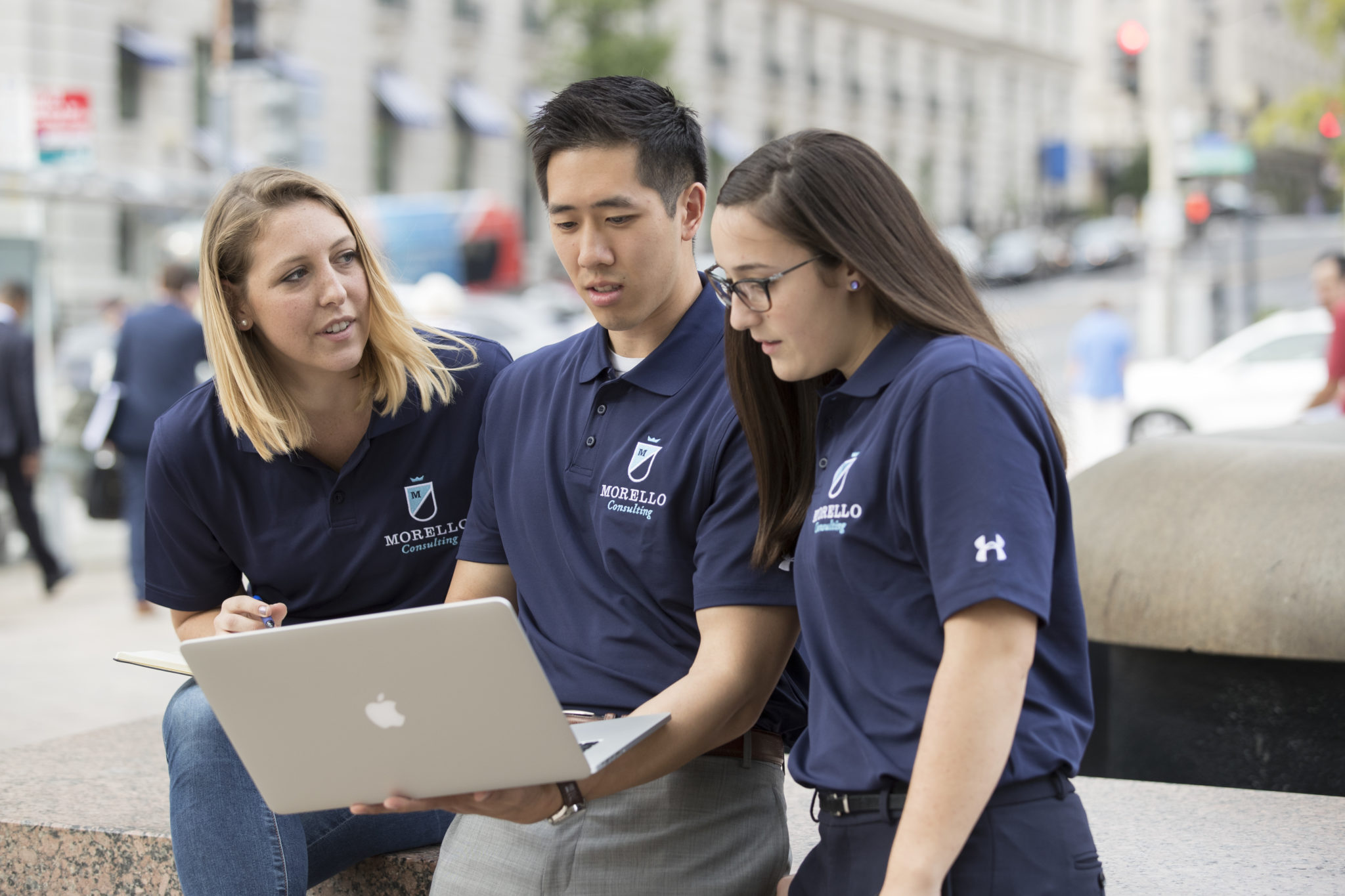 Three people looking at a laptop in a plaza wearing matching navy blue polos that say Morello Consulting.