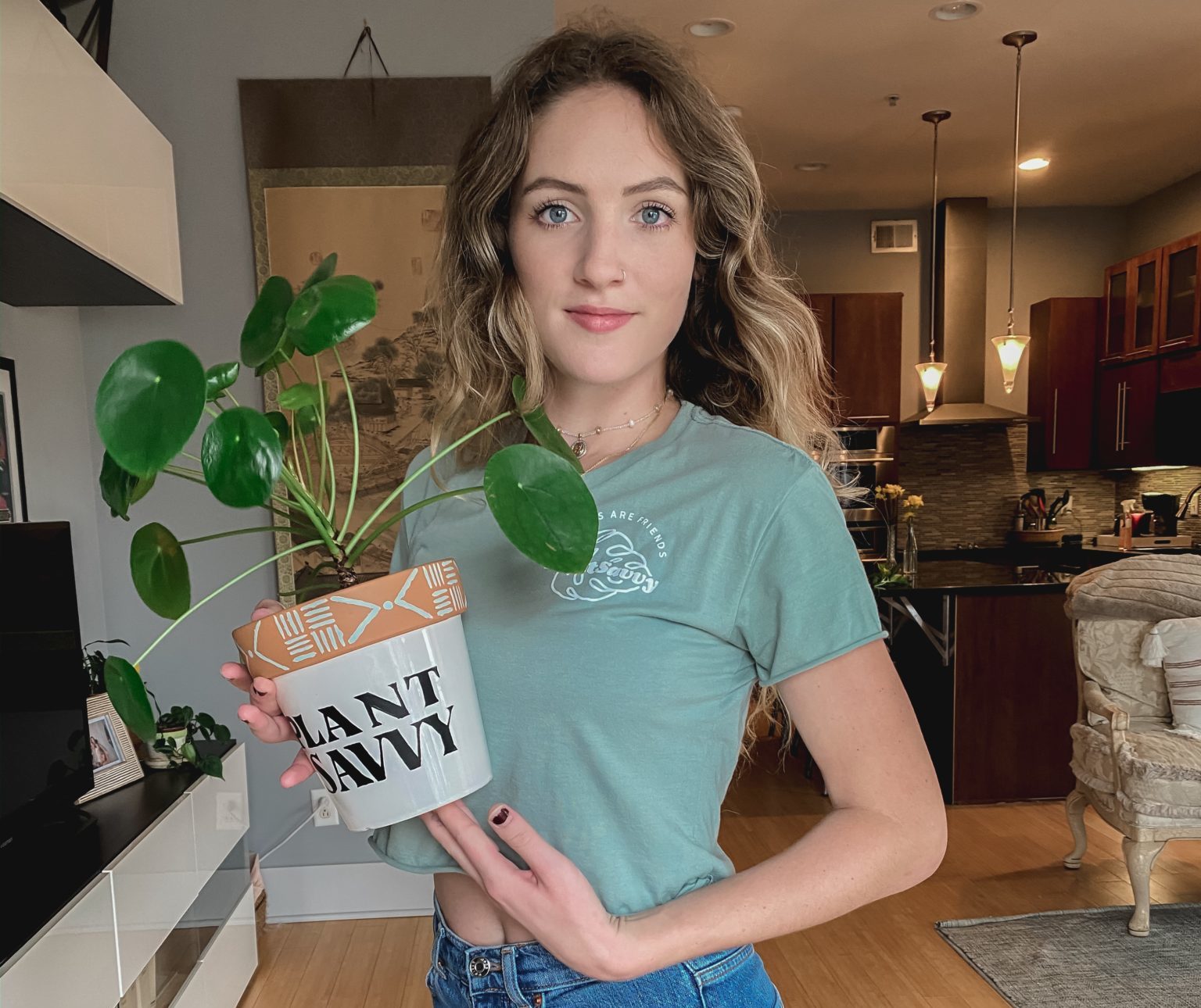 A woman faces camera in a light teal shirt, holding a plant in a pot bearing the text "PLANT SAVVY"